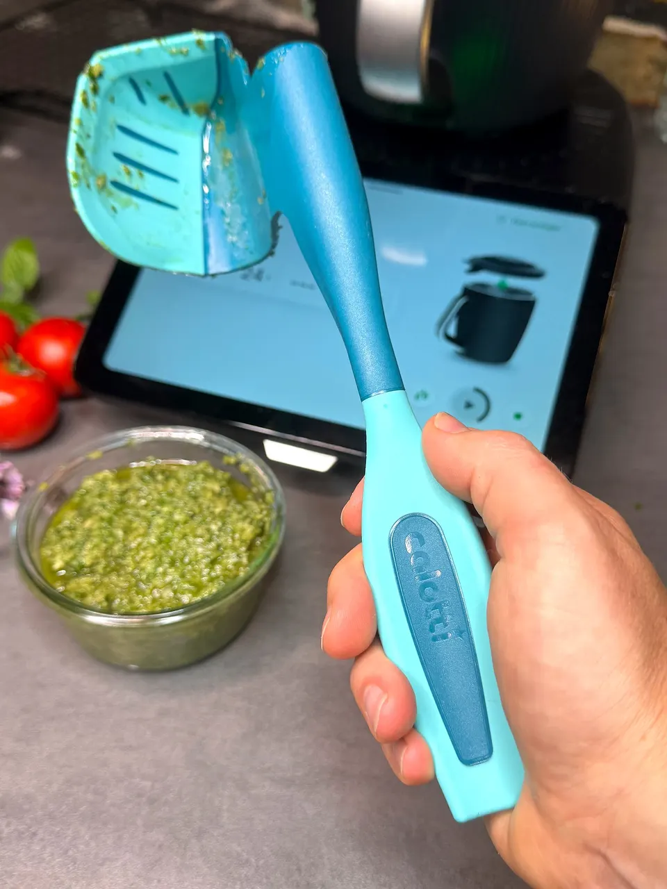 Calotti® Drehkellenspatel SILIKON+ in der Hand, dahinter Pesto und das Thermomix® TM7 Display